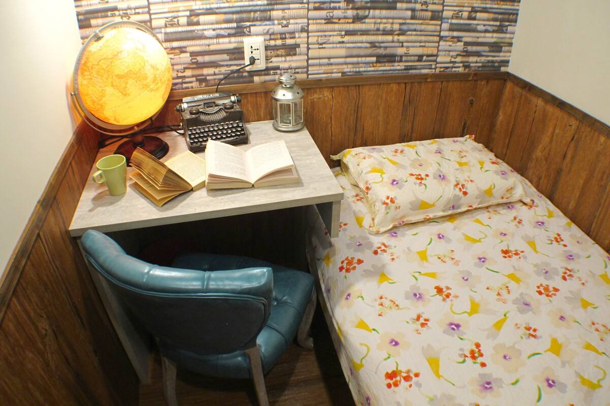 A small room with a desk, a chair, a bed, and a wall covered with newspapers.