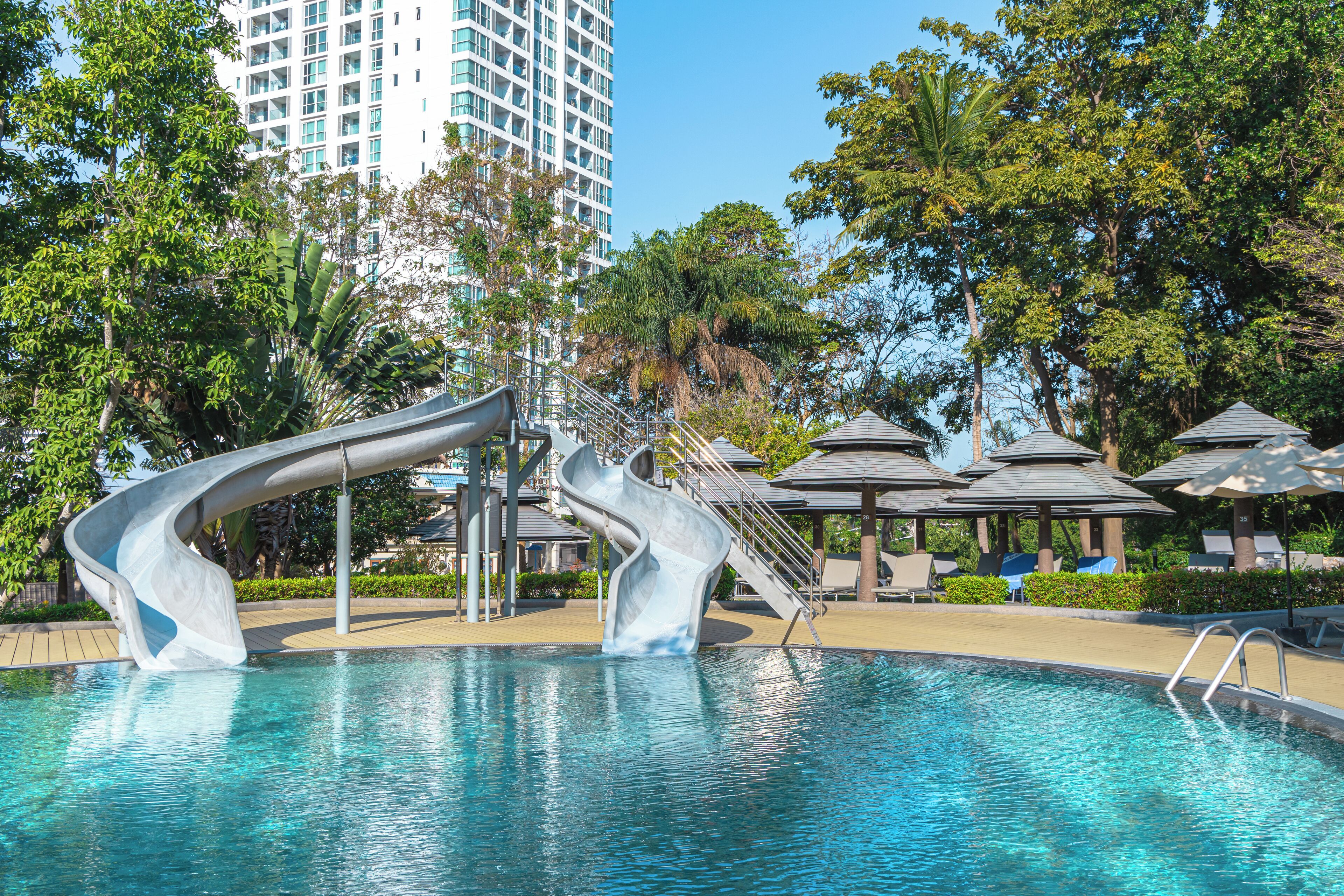 A swimming pool with a unique slide structure, surrounded by lounge areas and umbrellas, with a tall building in the background.
