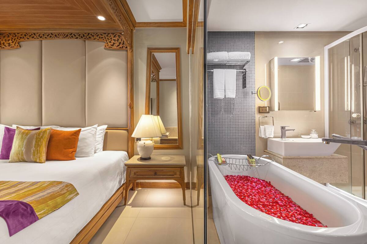 Une chambre d’hôtel avec un grand lit, une table de chevet avec une lampe, une salle de bain avec une baignoire remplie de pétales de rose et un espace douche.