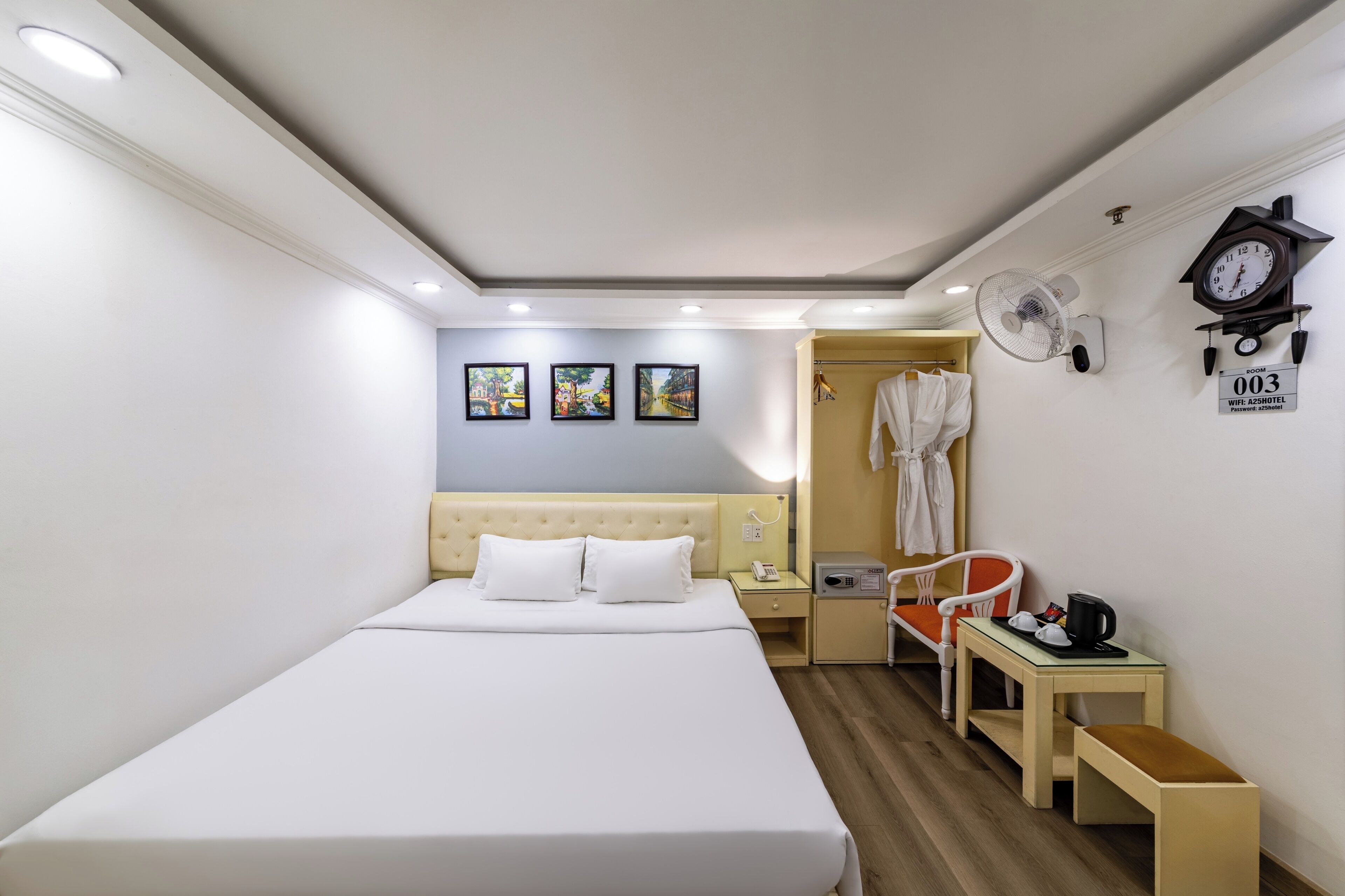 A hotel room with a bed, a bedside table with a lamp, a chair, a small table with a kettle, and a clock on the wall.