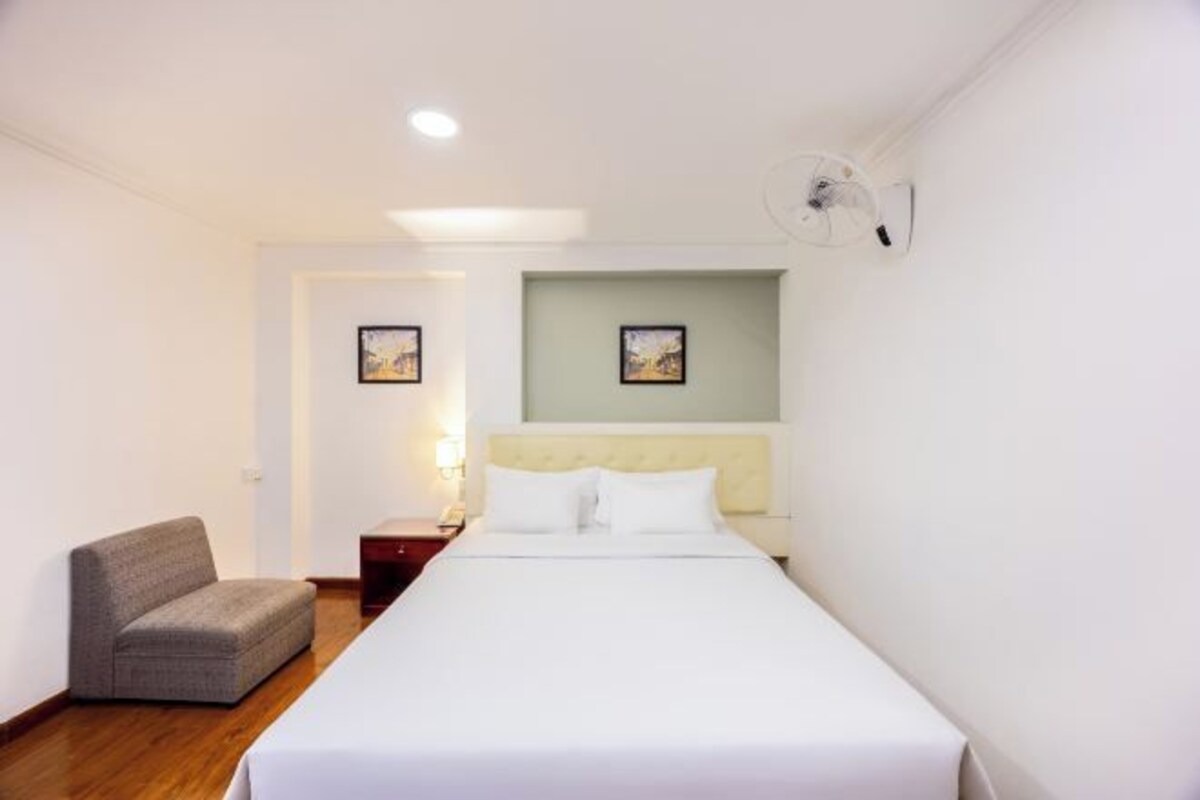 A hotel room with a single bed, a small armchair, a bedside table, and a wall-mounted fan.