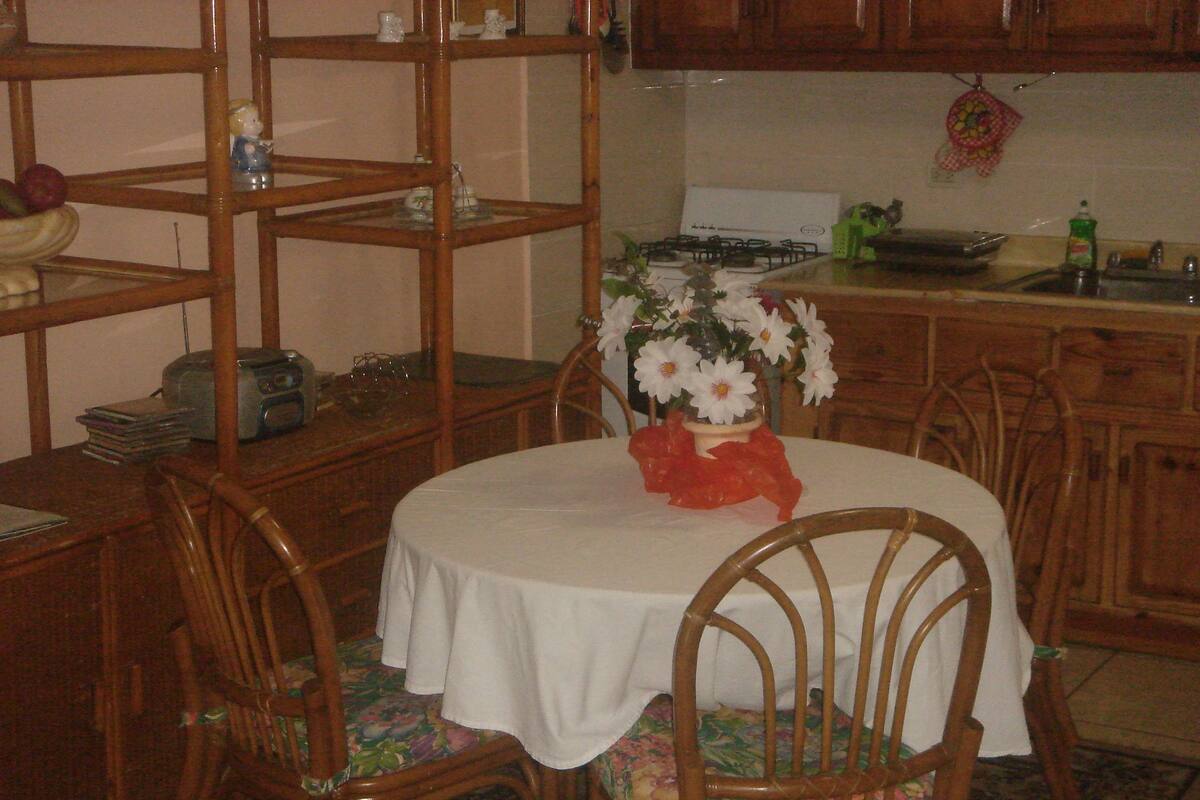 A dining area with a round table covered by a white tablecloth, surrounded by wooden chairs with floral upholstery. A vase with white flowers and a red ribbon is placed on the table.