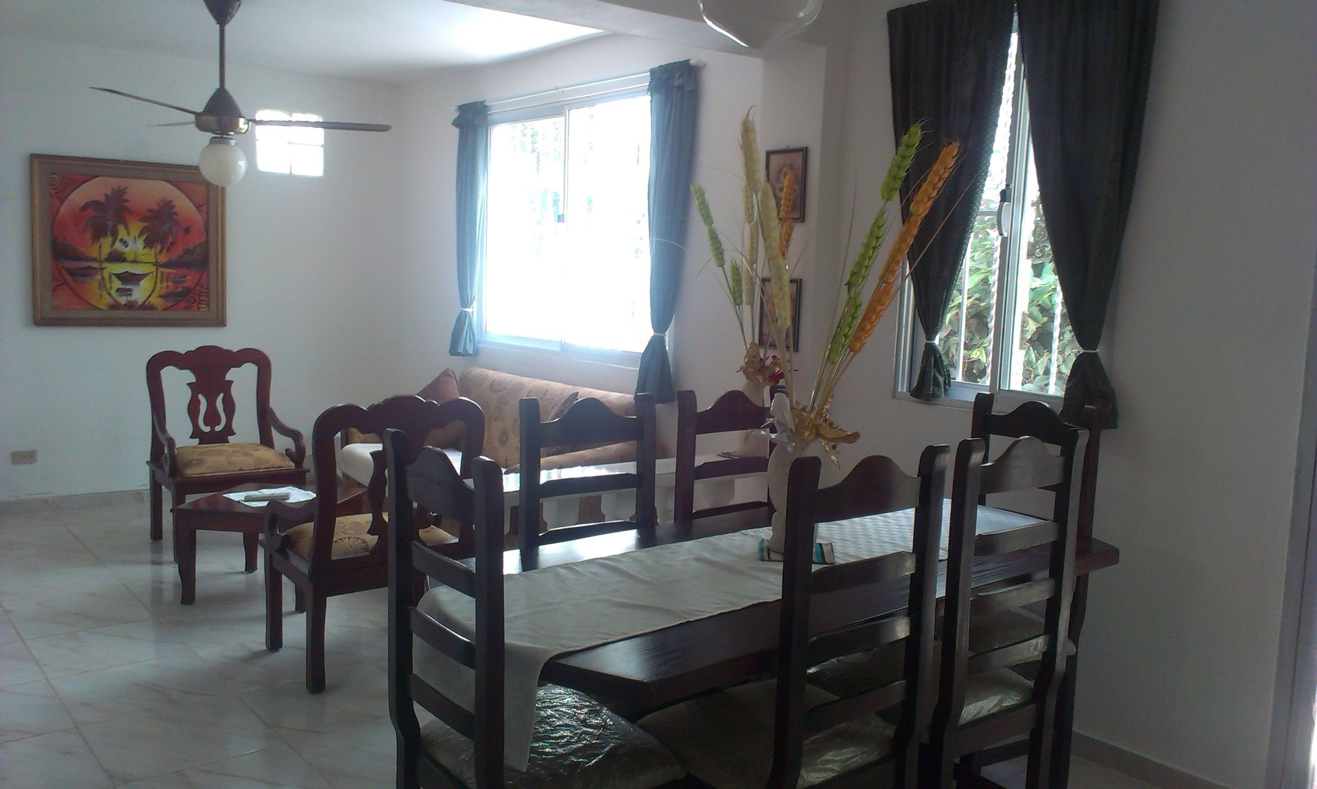 Un comedor con una mesa preparada para cuatro personas, rodeada de sillas de madera. Hay un ventilador de techo arriba, una ventana con cortinas y un cuadro en la pared.