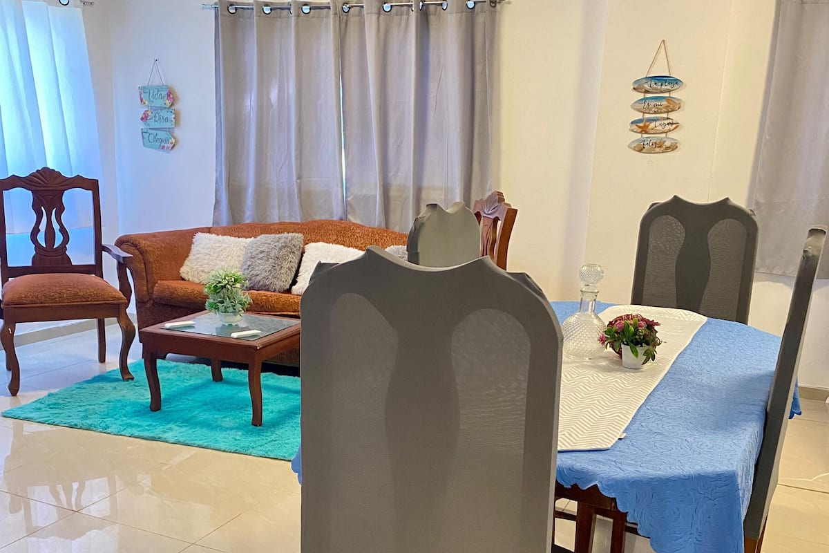 A living room with a sofa, chairs, a coffee table, and a dining table with a blue tablecloth.