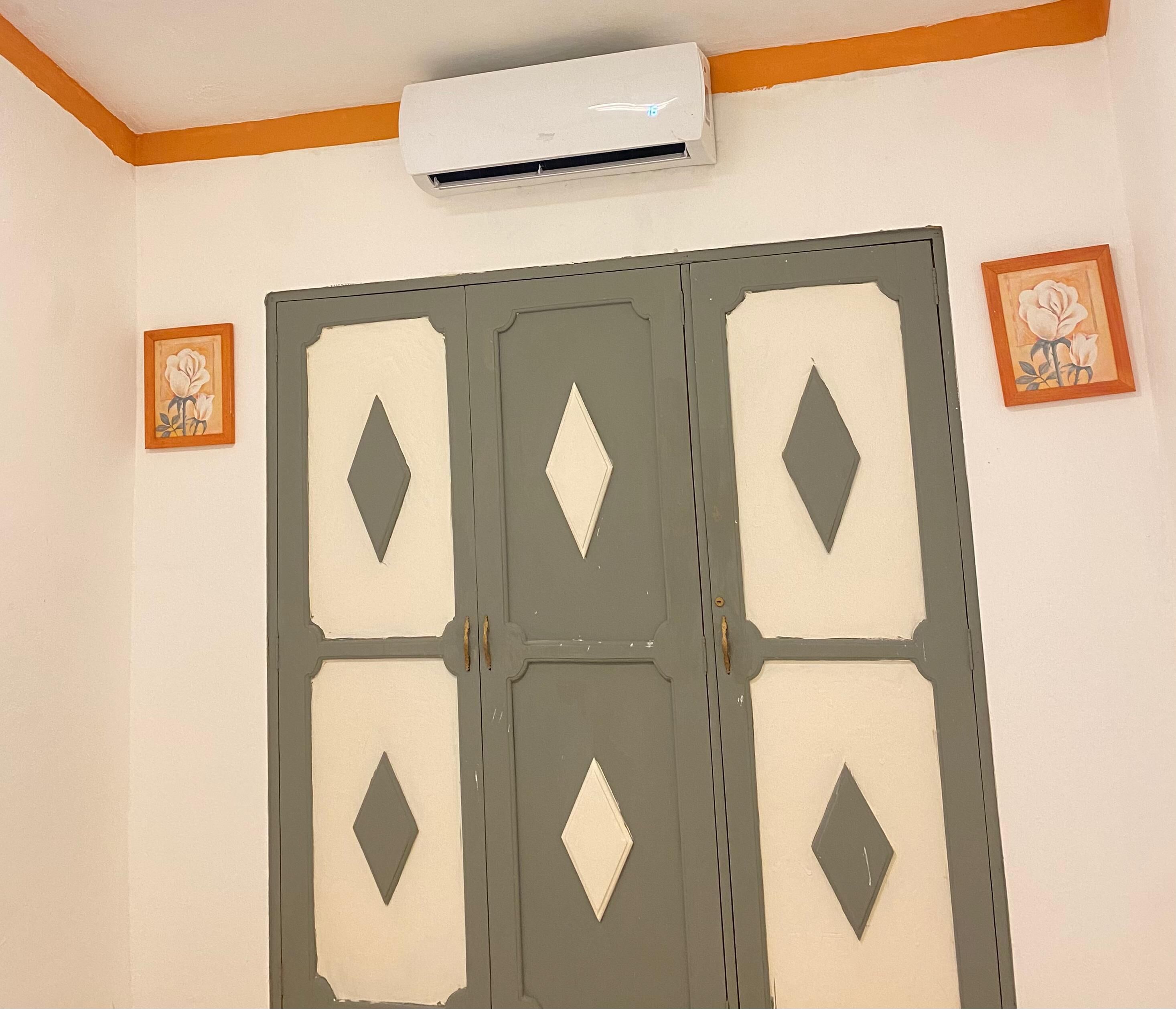 Een kamer met een grijze kast met diamantvormige uitsparingen, een airconditioningunit erboven en twee wanddecoraties met bloemen in lijsten.
