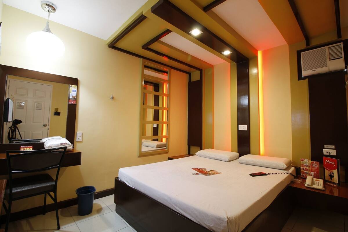 Une chambre d’hôtel équipée d’un lit, d’un bureau, d’une chaise, d’une poubelle, d’un miroir, d’une fenêtre et d’un climatiseur fixé au mur.