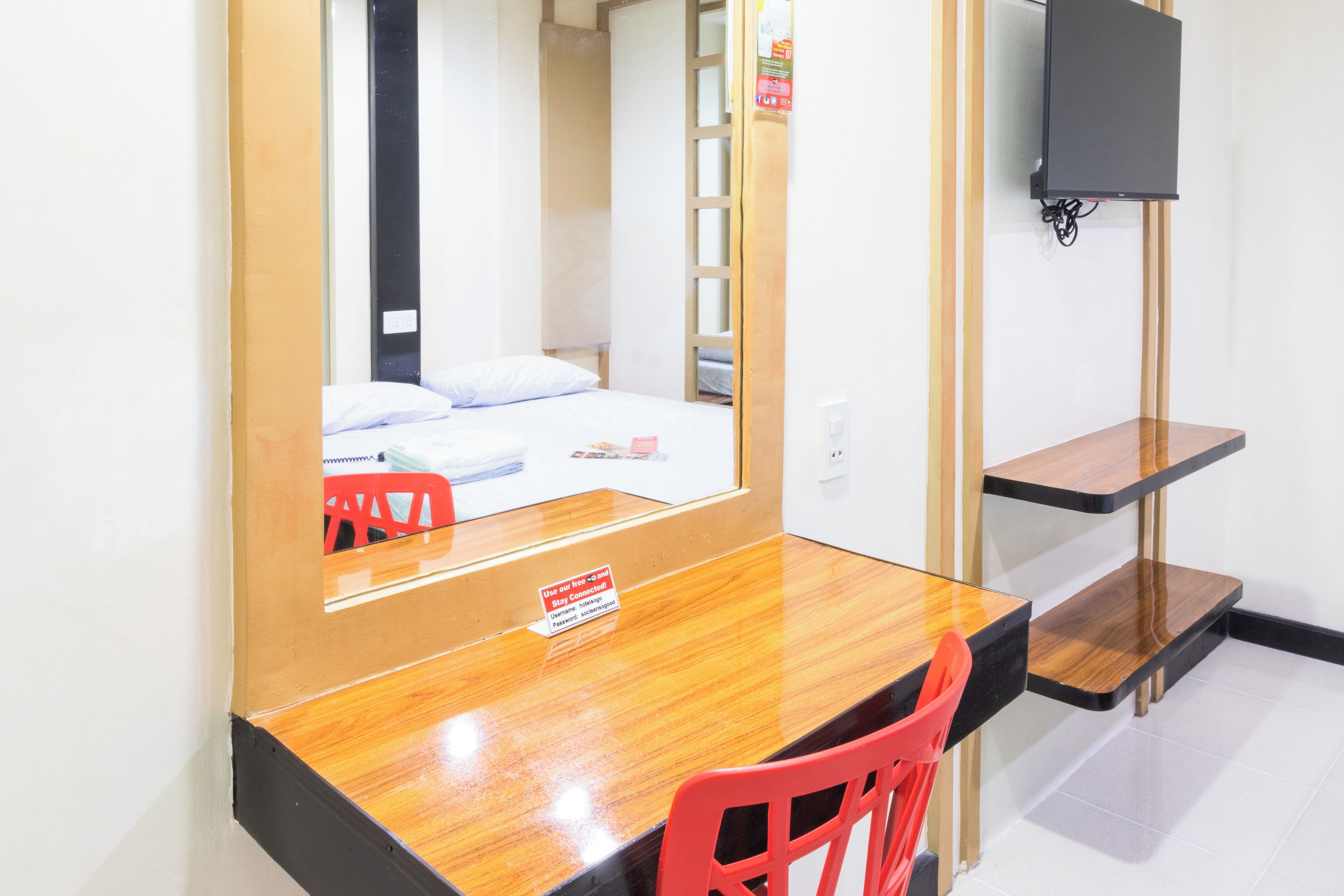 A hotel room with a wooden desk, a red chair, a bed with white bedding, a TV mounted on the wall, and a small shelf.
