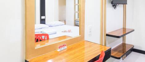 A hotel room with a wooden desk, a red chair, a bed with white bedding, a TV mounted on the wall, and a small shelf.
