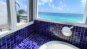 A bathroom with blue tiles, a white bathtub, and a window offering a view of the ocean and a rocky outcrop. - Rockhand Homestay (Hengchun)