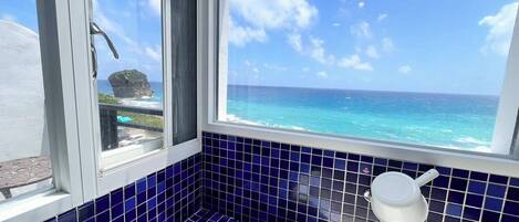 A bathroom with blue tiles, a white bathtub, and a window offering a view of the ocean and a rocky outcrop.