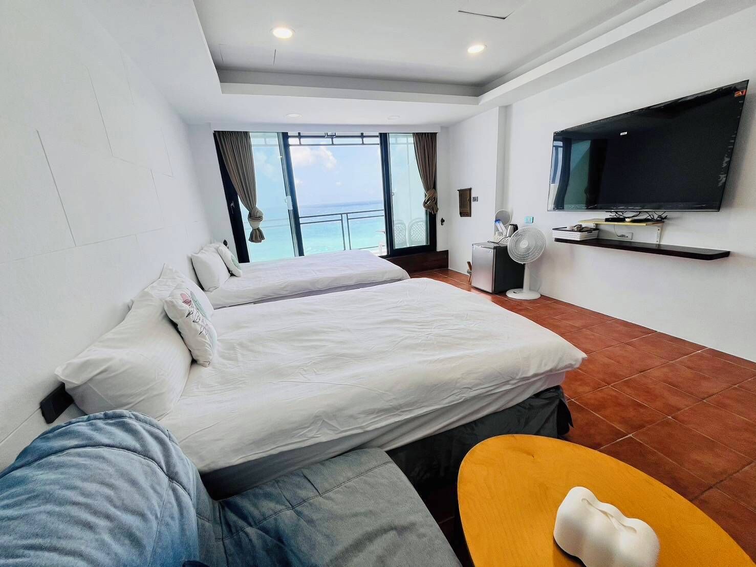 A hotel room with two beds, a TV, a fan, and a view of the sea.