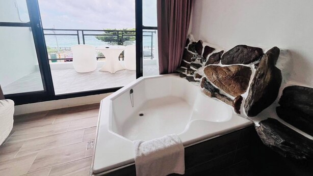 A modern bathroom with a freestanding bathtub, a stone accent wall, and a view of the outdoor terrace with white furniture. - Rockhand Homestay (Hengchun)