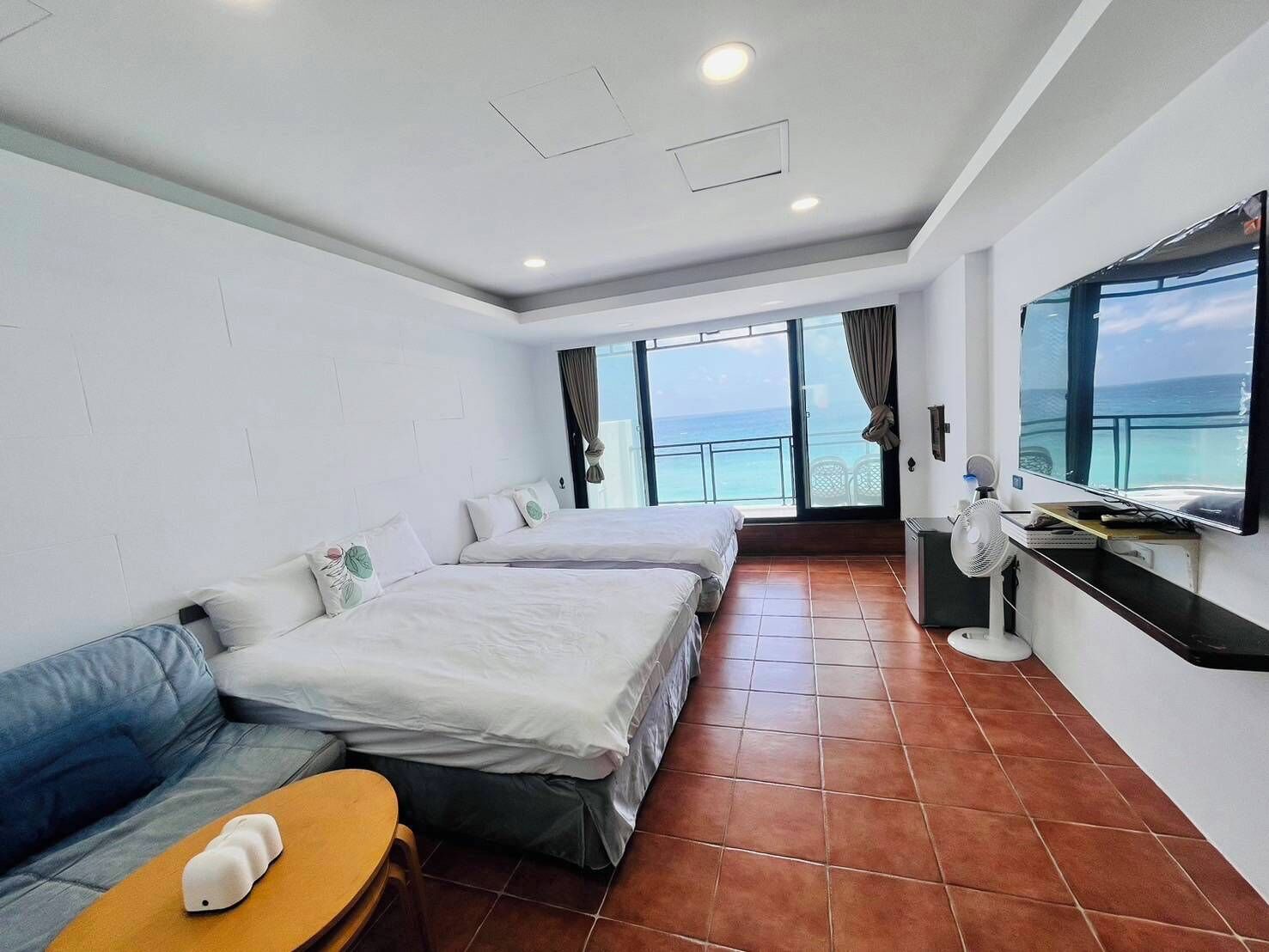 A hotel room with two beds, a sofa, a small table, a TV, and a view of the sea.