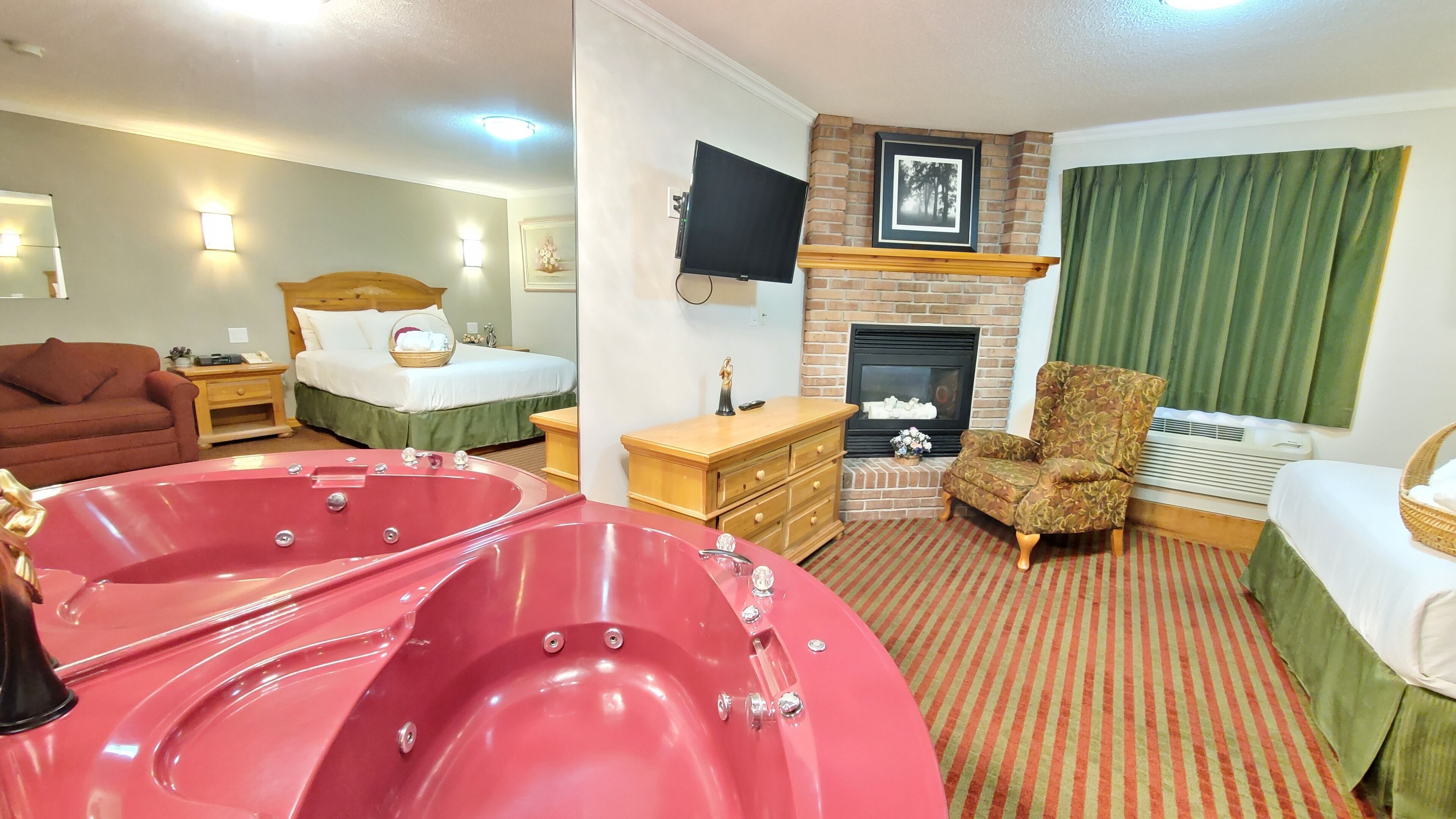 Hotelkamer met een groot rood bubbelbad, een bed met groen beddengoed, een houten dressoir, een televisie, een open haard en een groen gordijn.
