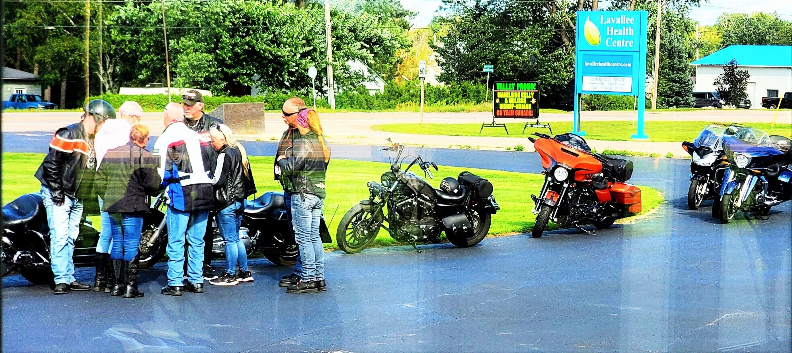Un grupo de personas de pie cerca de motocicletas en un estacionamiento, con un letrero del Centro de Salud Layallee al fondo.