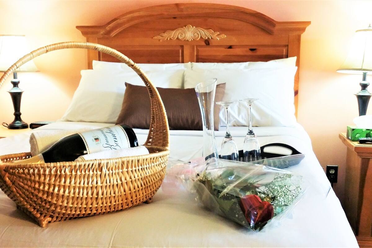 A neatly made bed with a basket of wine, a bouquet of flowers, and a bottle of champagne.