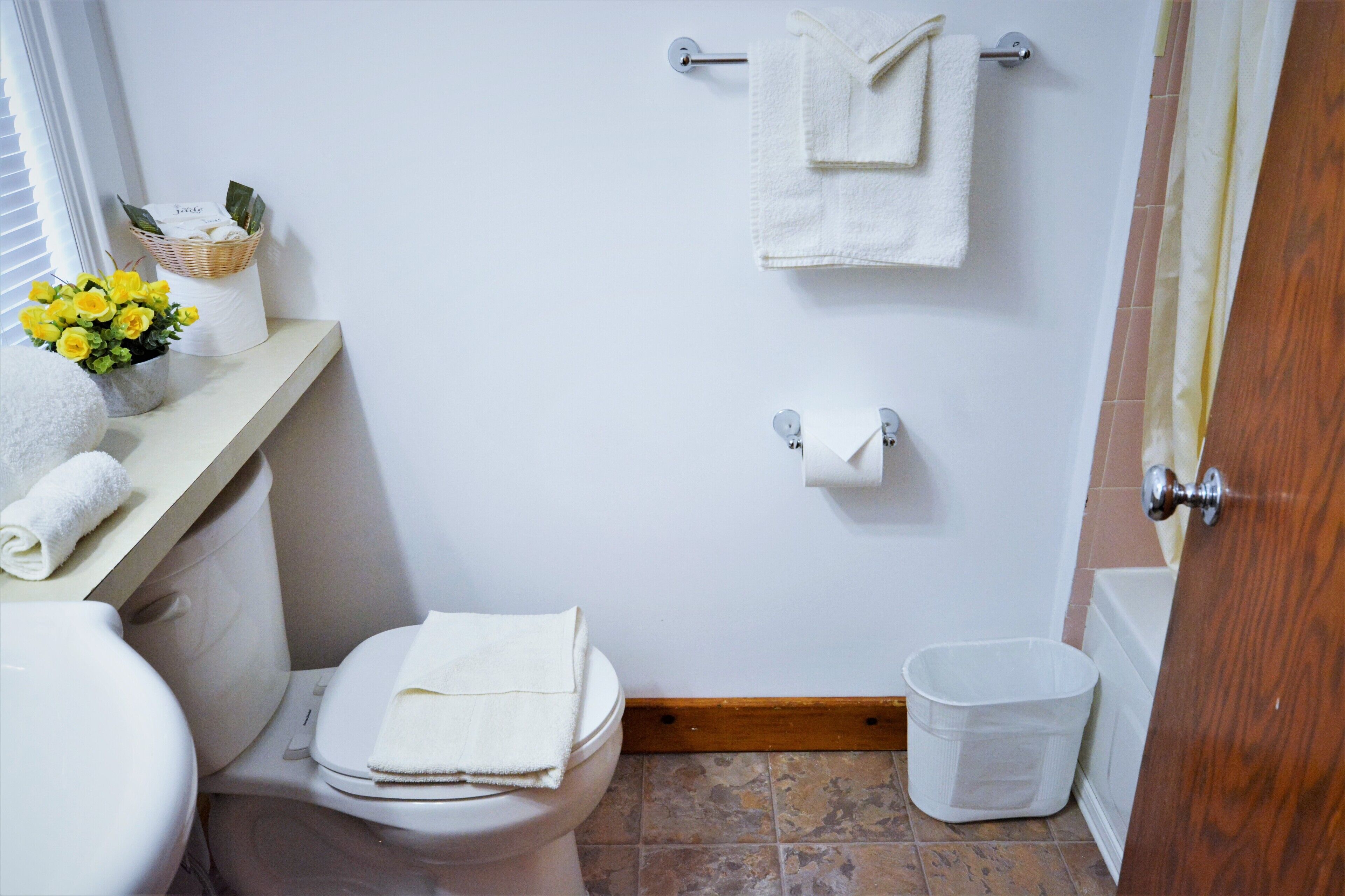 Een kleine badkamer met een toilet, een wastafel, een handdoekrek met twee handdoeken, een mand met bloemen en een prullenbak.