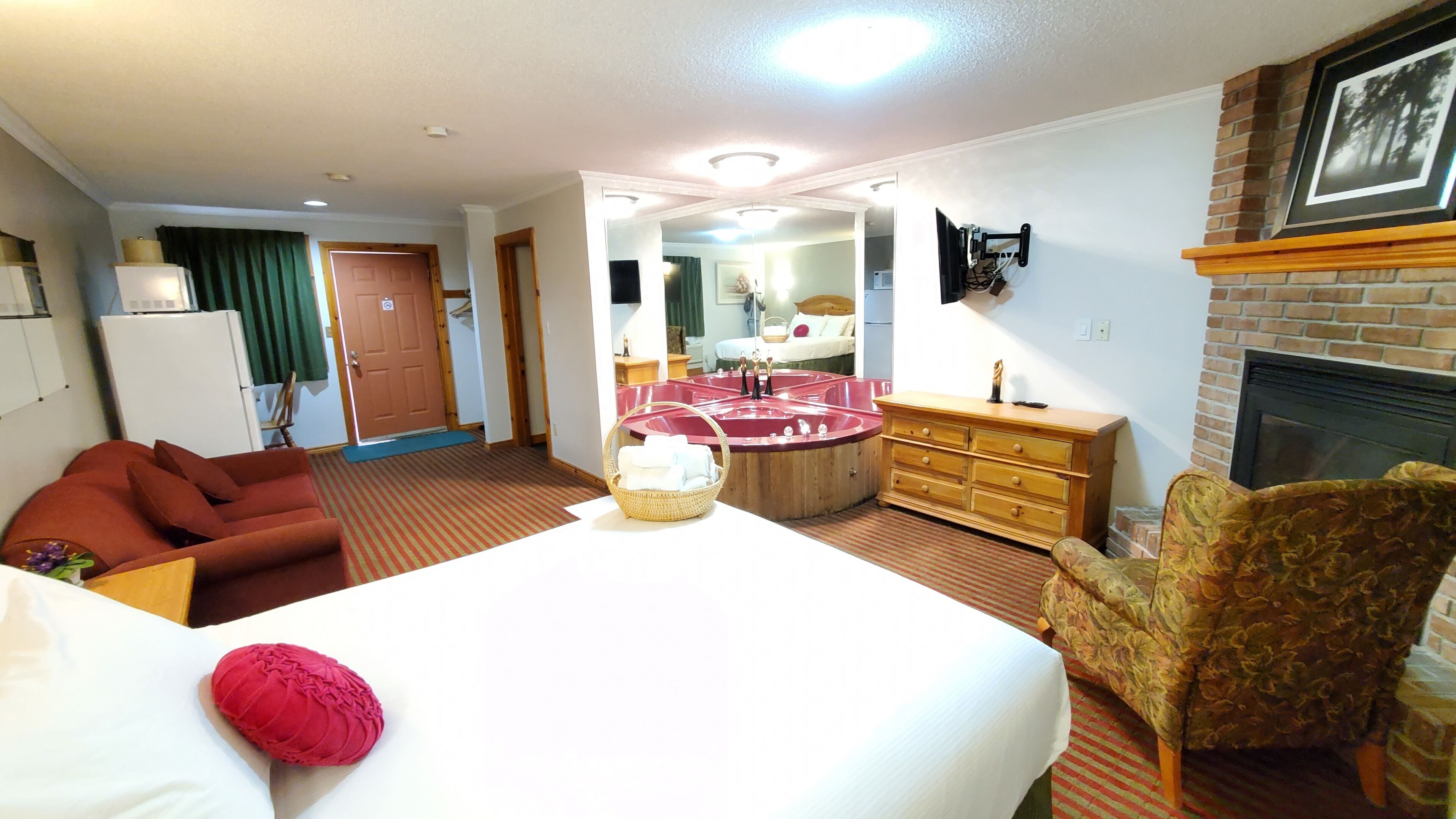 Une chambre d’hôtel avec un lit, un canapé rouge, une commode en bois, une cheminée et un jacuzzi rond.