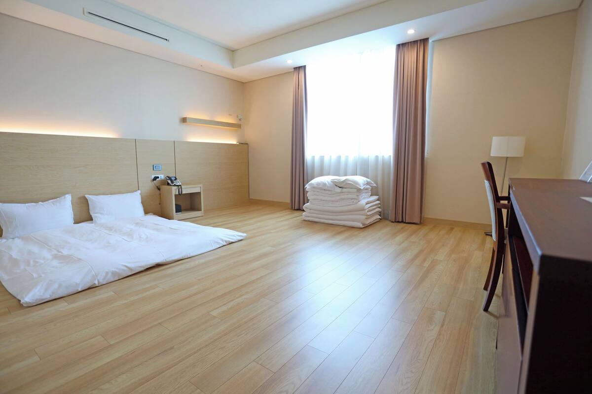 A hotel room with a large bed, a desk with a lamp, a chair, and a window with curtains.