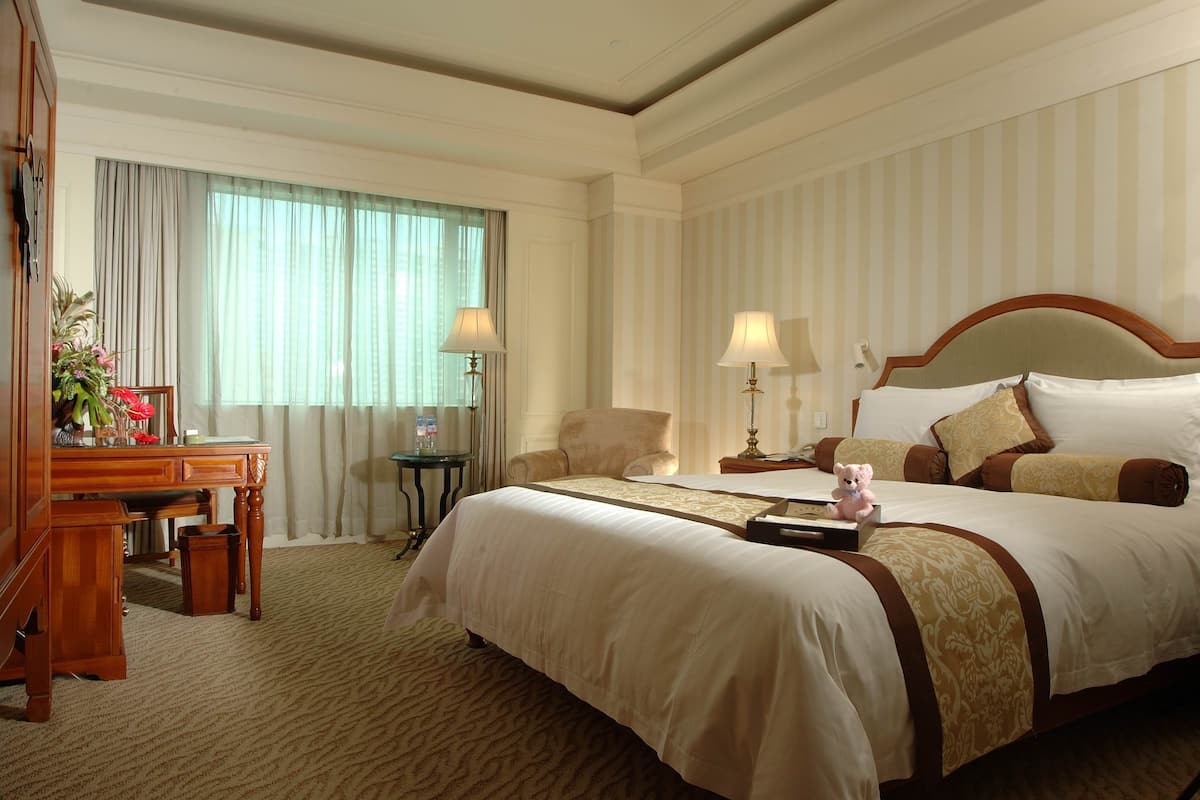 Een hotelkamer met een groot bed, twee nachtkastjes met lampen, een houten bureau met stoel, en een decoratieve vaas.
