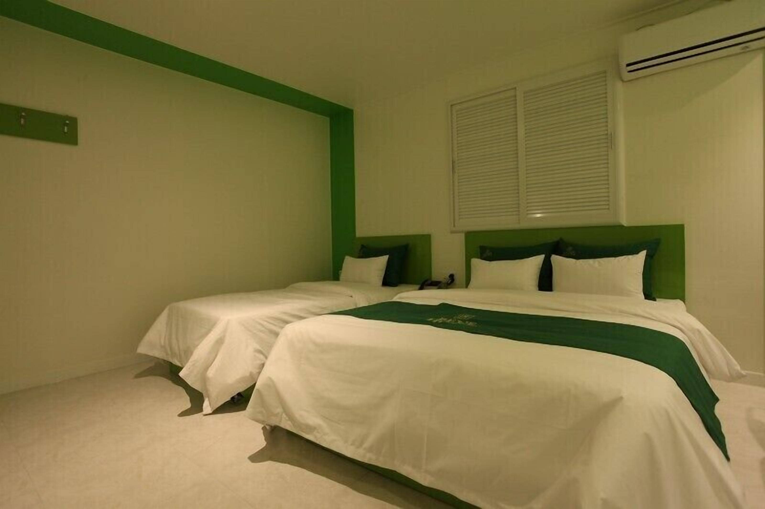 Ein Zimmer mit zwei Betten, jeweils mit weißer Bettwäsche und grünen Kopfteilen. Ein Fenster mit Jalousien, eine Klimaanlage an der Wand und ein kleiner Tisch mit einer Tischdecke.