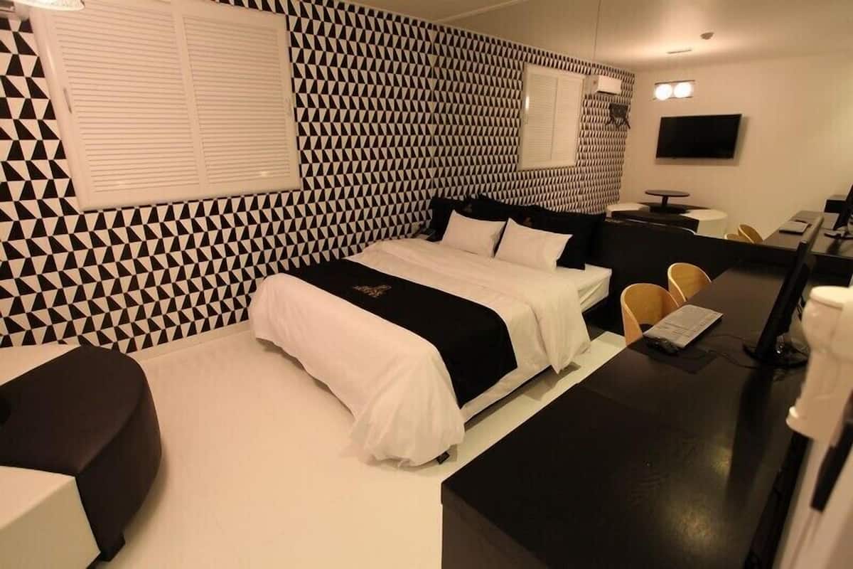 A modern hotel room with a geometric patterned wall, a large bed with white and black bedding, a desk with a chair, and a flat-screen TV.