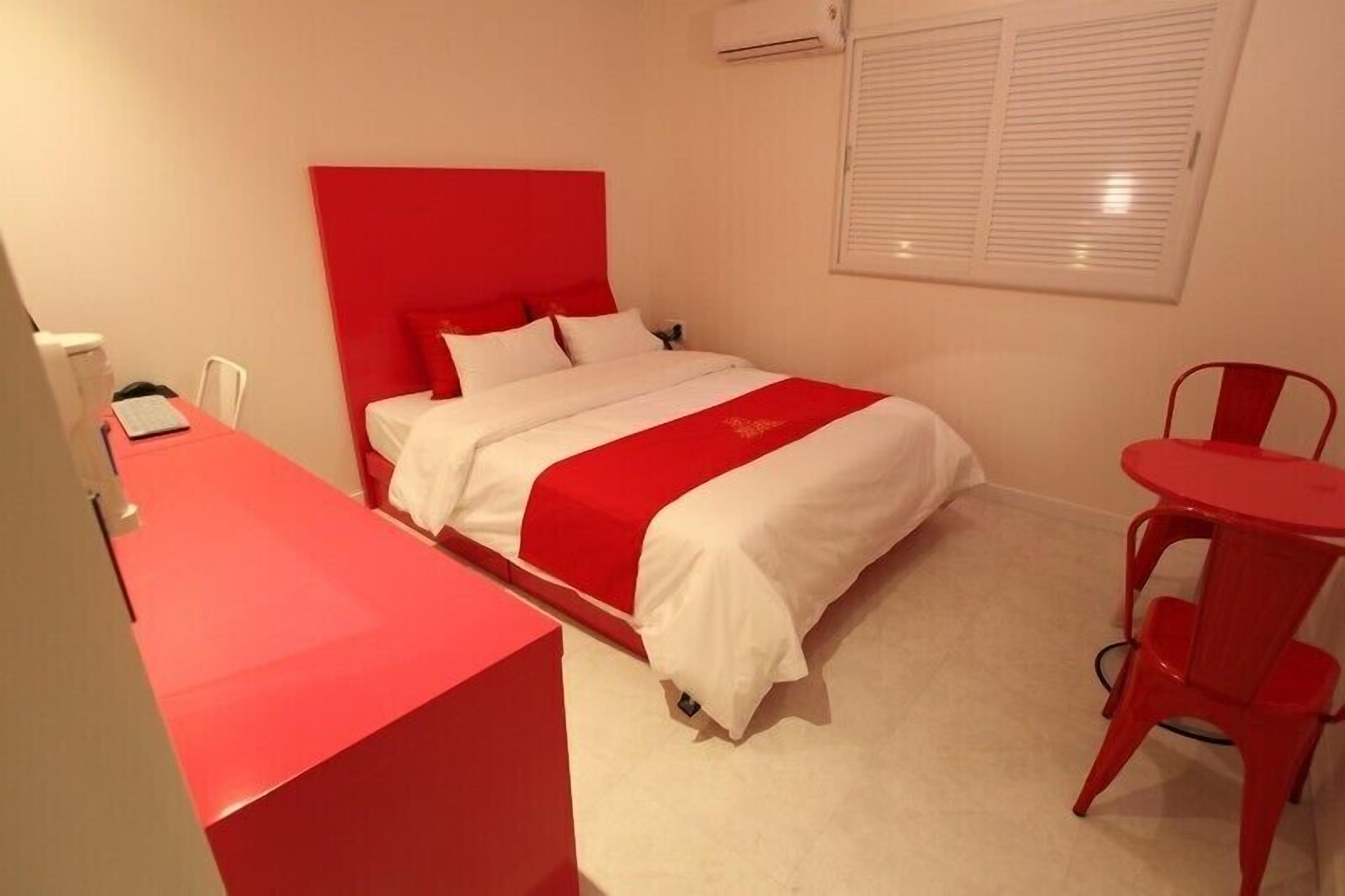 Una habitación de hotel con una cama roja y blanca, un escritorio pequeño con una silla, y una butaca roja.