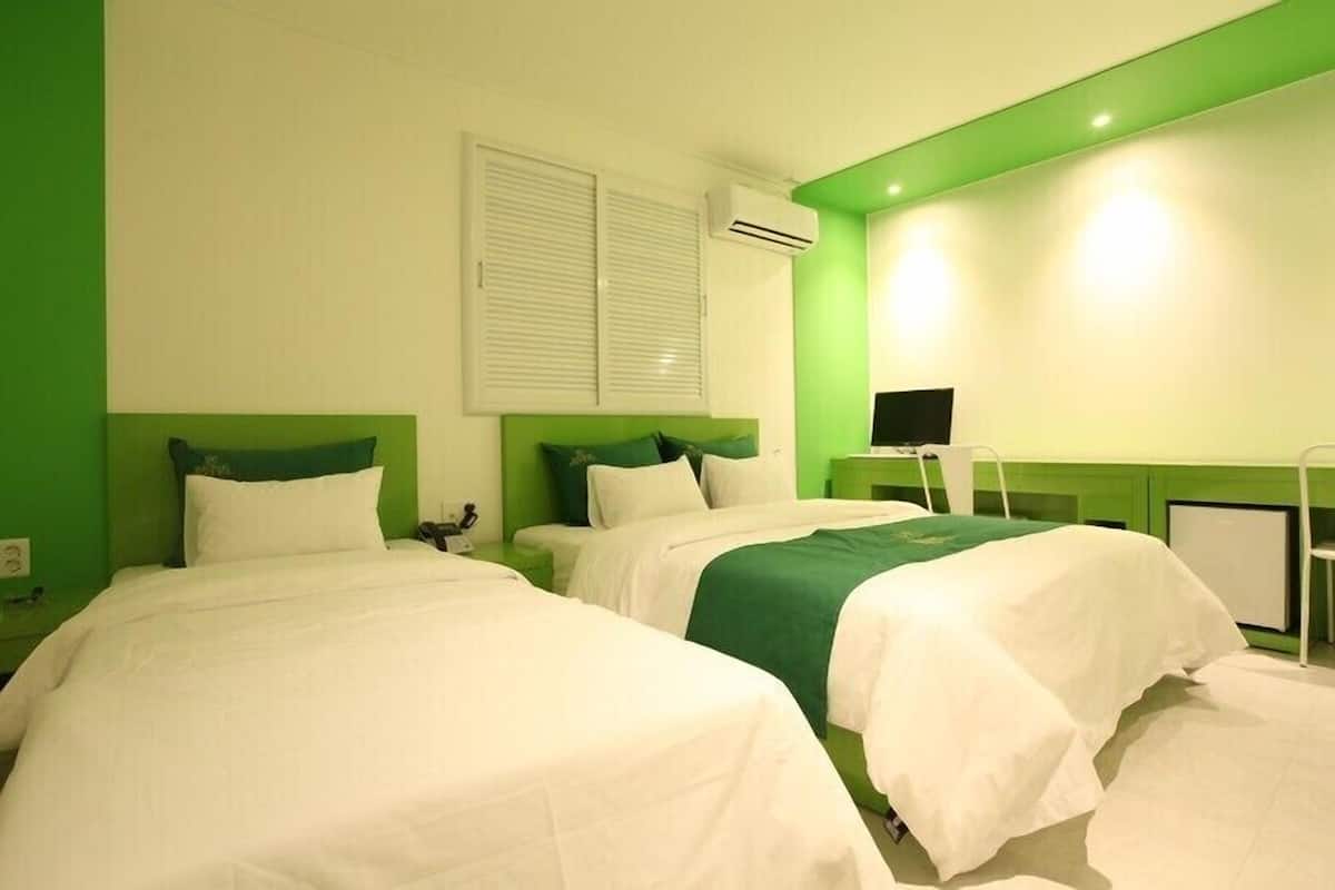 A hotel room with two beds, green walls, white bedding, an air conditioning unit, and a small desk with a chair.