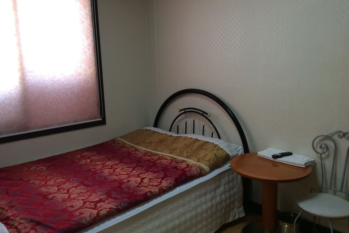 A small, single-bed room with a red bedspread, a wooden side table, a white chair, and a window with a curtain.