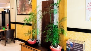 A reception area with a wooden reception desk, two potted plants, a large framed picture on the wall, and a promotional stand. - Hotel Sogo Novaliches (Quezon City)