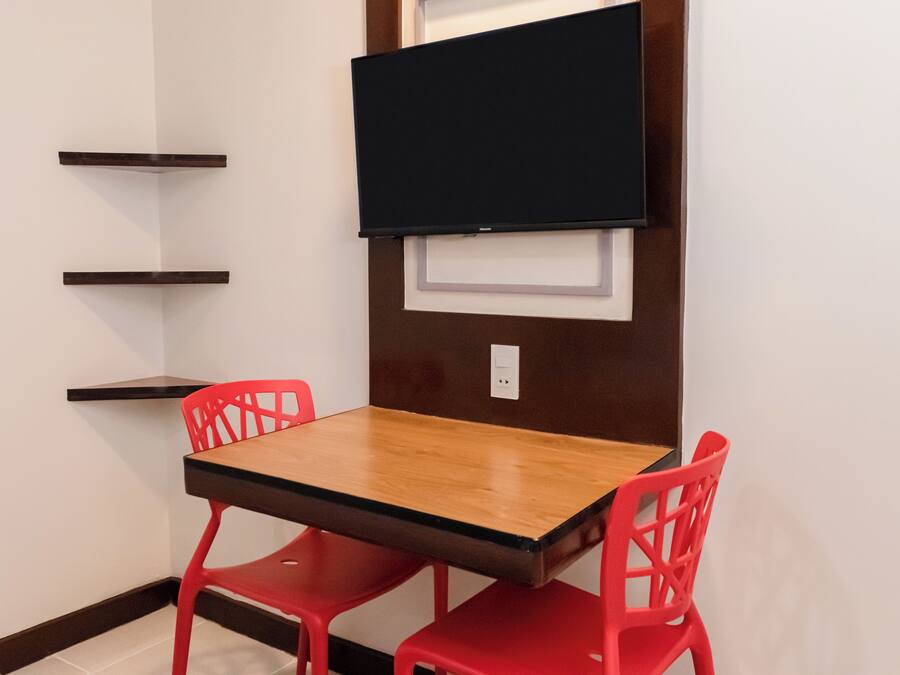 A small room with a wooden table and two red chairs, a black TV mounted on the wall, and two wooden shelves.