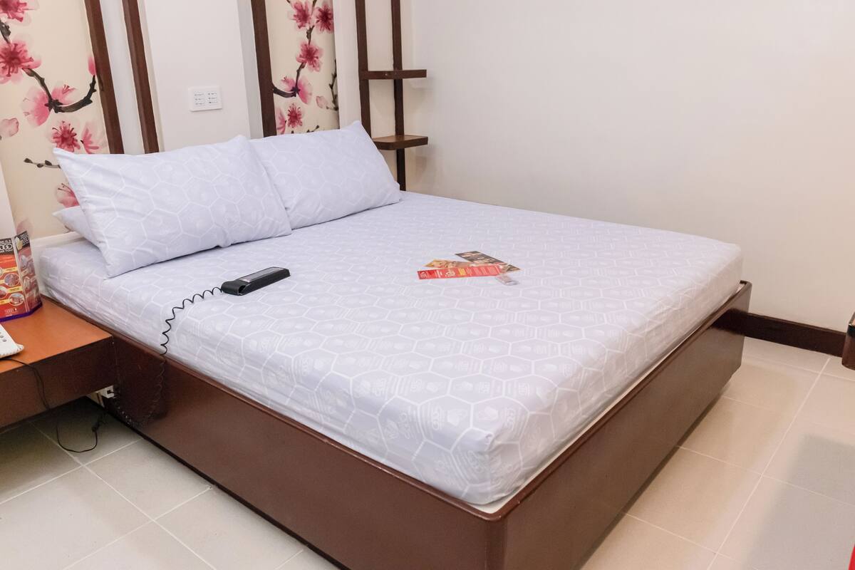 A neatly made bed with a white mattress and a geometric-patterned duvet, a bedside table with a phone and a book, and a wall with a floral wallpaper design.