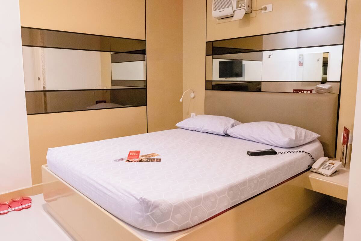 A hotel room with a bed, a bedside phone, a remote control, and a mirror on the wall.
