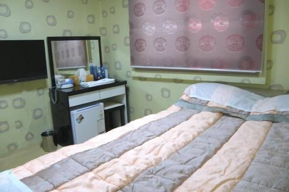 A bedroom with a bed, a bedside table with a mirror, a small desk, and a window with a patterned curtain.