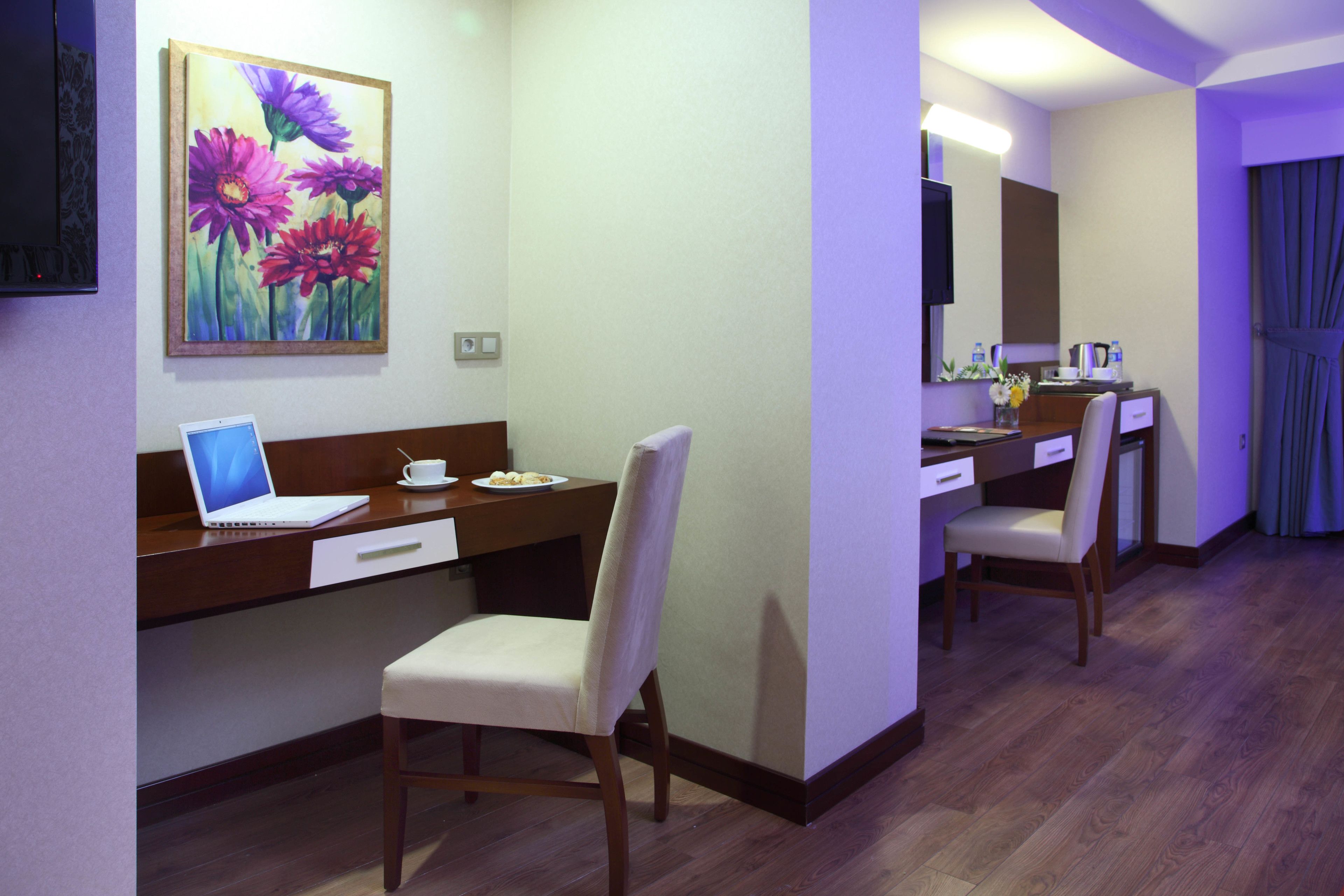 A hotel room with a desk, laptop, chair, and a painting of flowers on the wall.