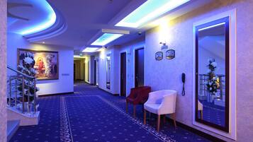 A hotel hallway with a blue carpet, a staircase with a metal railing, a red chair, a white chair, and a large mirror on the wall.