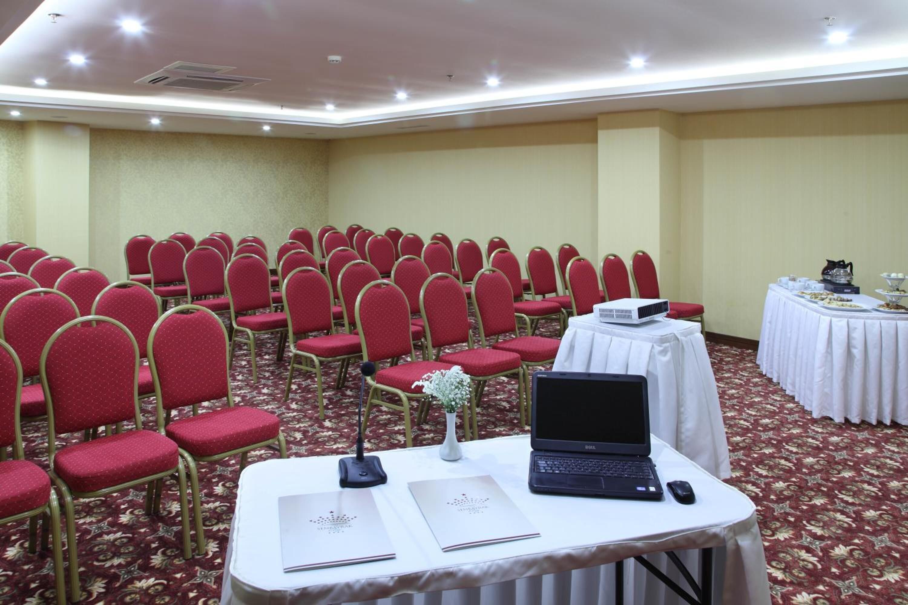 Une salle de conférence avec des rangées de chaises rouges, une table blanche sur laquelle se trouve un ordinateur portable, et une table de buffet en arrière-plan.