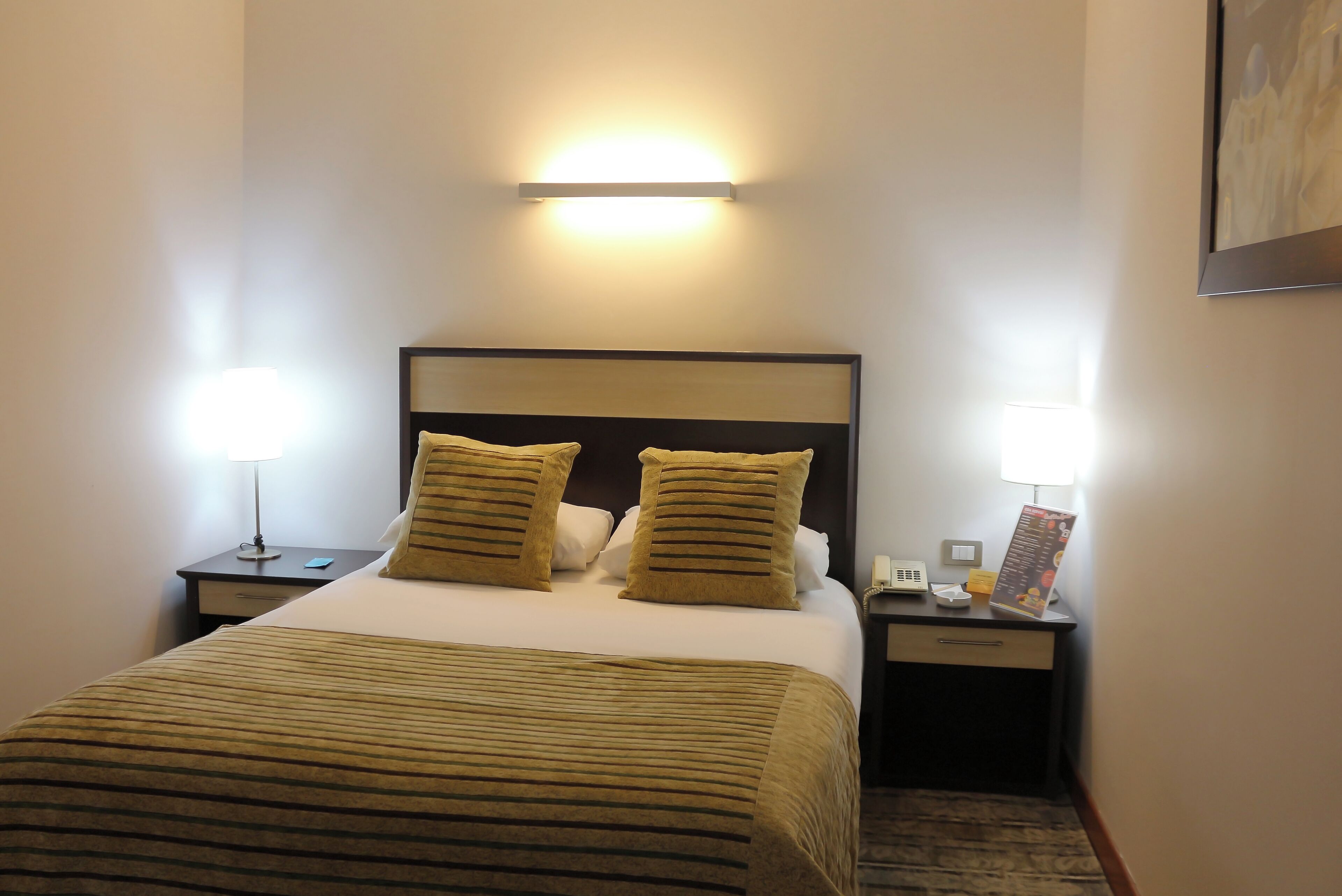 Una habitación de hotel con una cama, dos lámparas de noche, una mesita de noche, un teléfono y un cuadro en la pared.