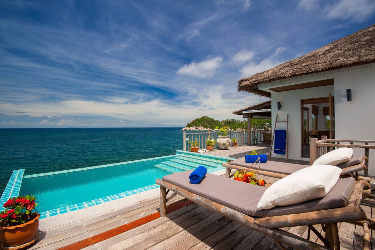 An infinity pool with a wooden deck, lounge chairs, and a thatched-roof building overlooking the ocean.