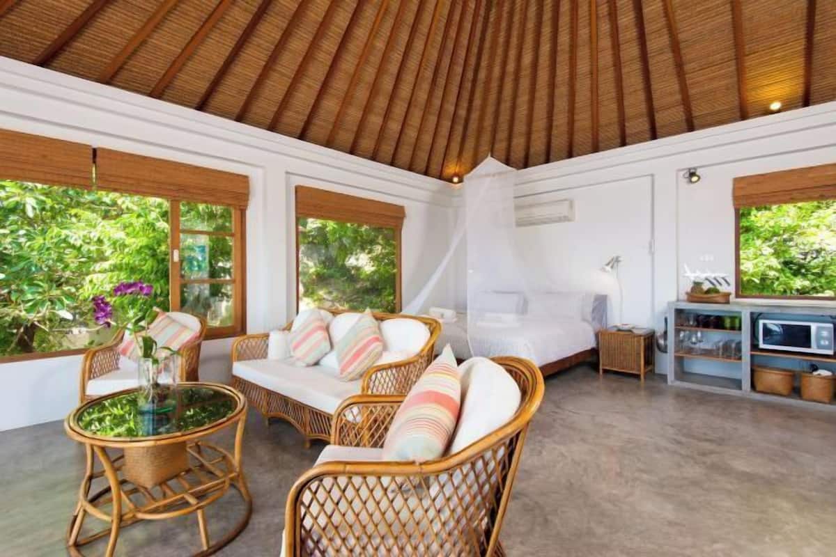 A room with wicker furniture, a bed with a mosquito net, and a kitchenette.