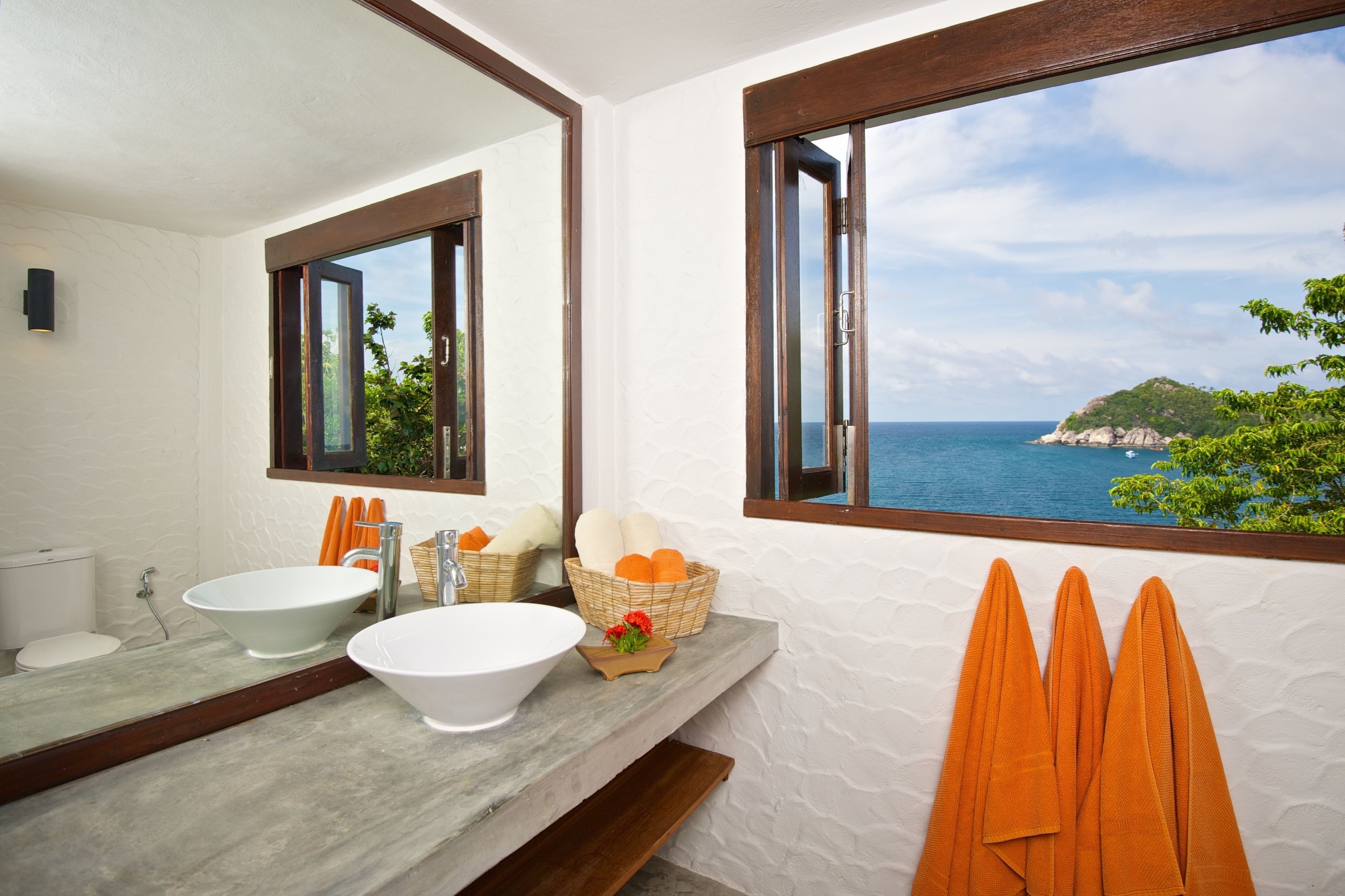 A bathroom with a large mirror, two sinks, a window with a view of the sea, and orange towels hanging on the wall.