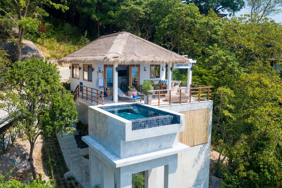 A modern villa with a rooftop infinity pool, a thatched-roof structure, and lush greenery surrounding the property.