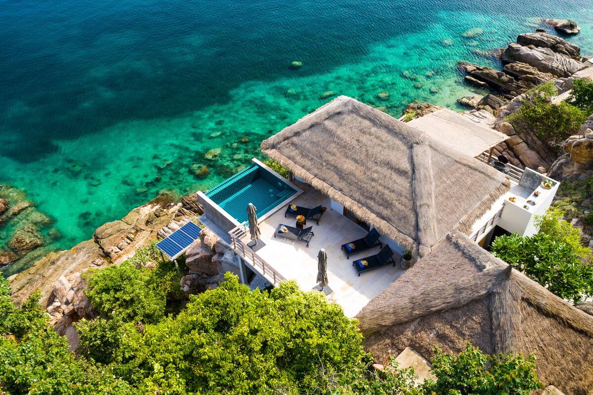 A luxurious villa with a private infinity pool overlooking a clear blue sea, surrounded by rocky terrain and lush greenery.