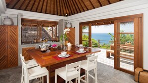 A dining area with a wooden table set for four, a view of the ocean, and a thatched roof ceiling.