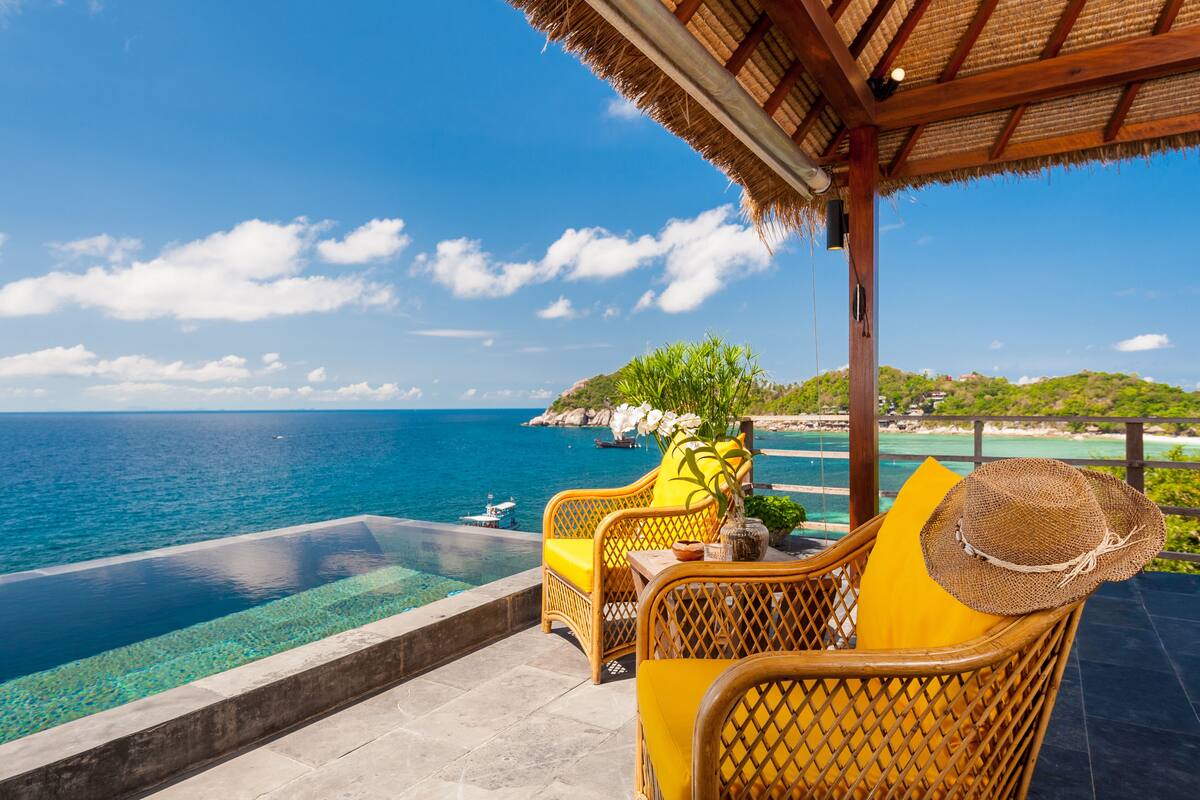 A seaside terrace with wicker furniture, a clear pool, and a view of the ocean and distant land.