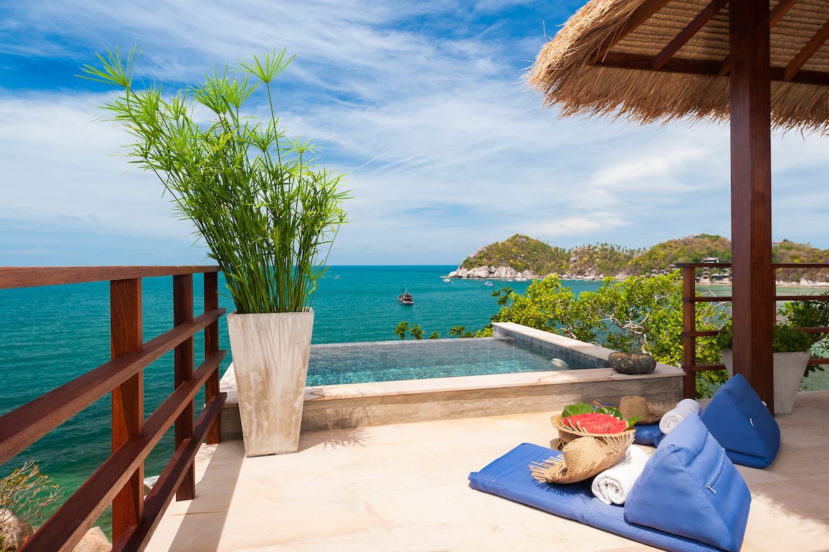 A balcony with a view of the sea, a pool, and a seating area with cushions and a basket of fruit.