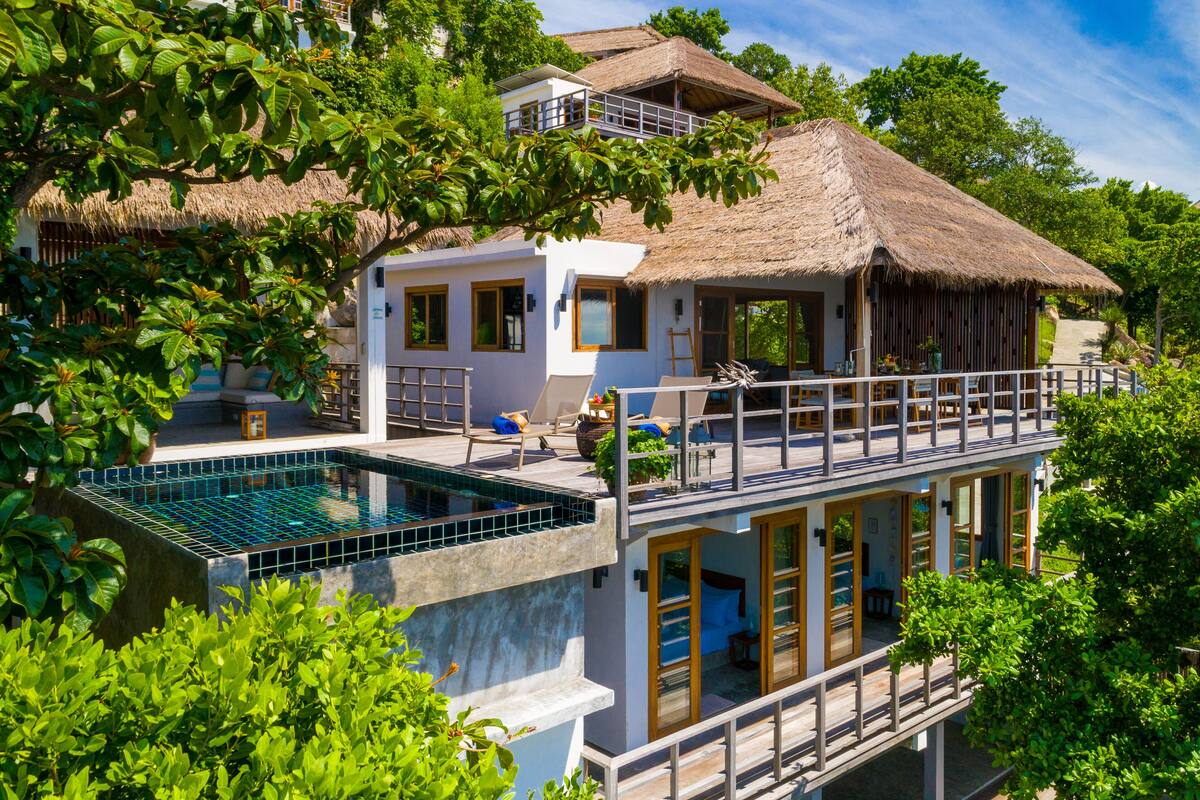 A two-story house with a swimming pool, a balcony, and a thatched roof.