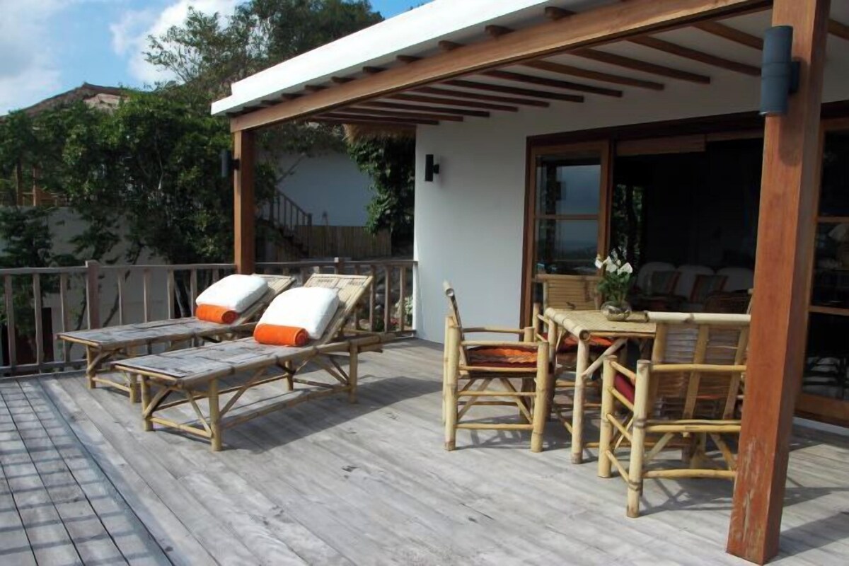 A wooden deck with lounge chairs and a table, a pergola, and a view of a building with a glass door.