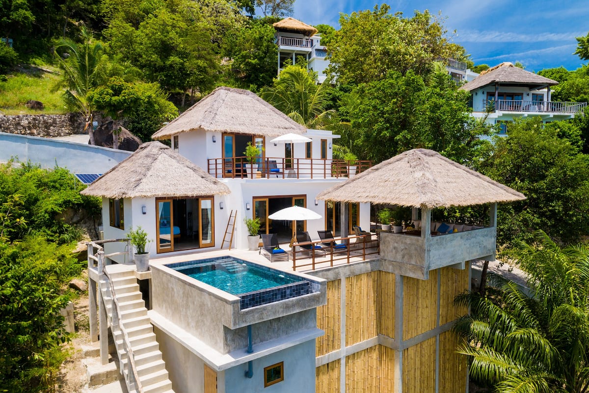 A two-story villa with a swimming pool, a balcony, and a thatched roof.