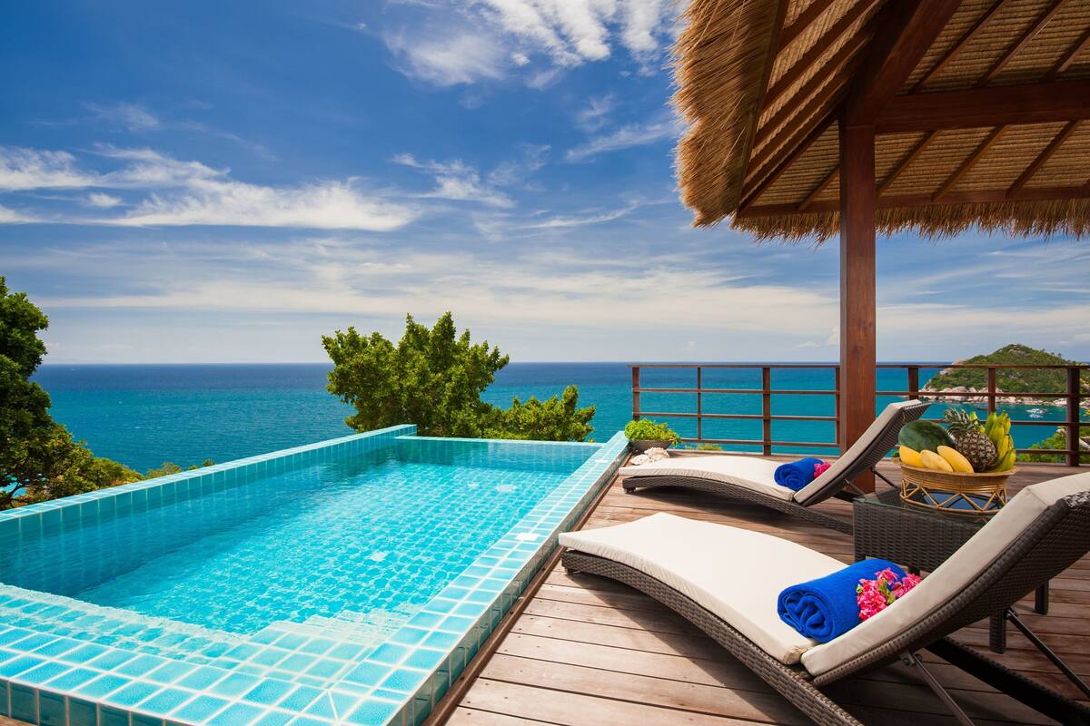 A swimming pool with a view of the ocean, lounge chairs, and a thatched roof structure.